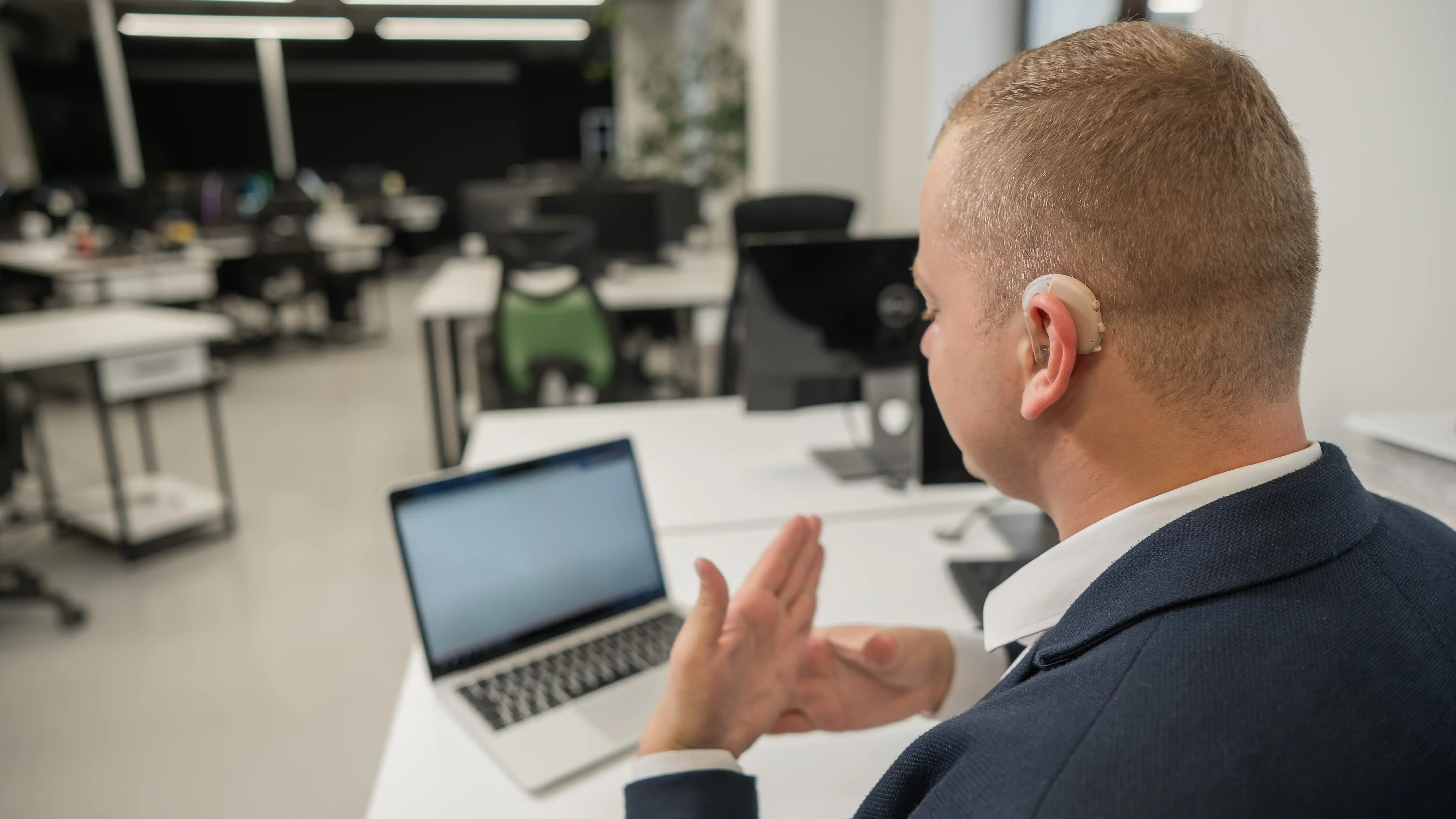 An office worker wearing a hearing aid signing or gesturing during a video call on a laptop, representing an employee protected against retaliation for requesting accommodations under California FEHA