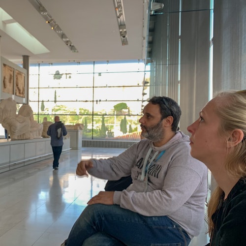 两个人坐在现代博物馆内的长椅上，欣赏着雕塑。阳光透过他们身后的大窗户洒进来。