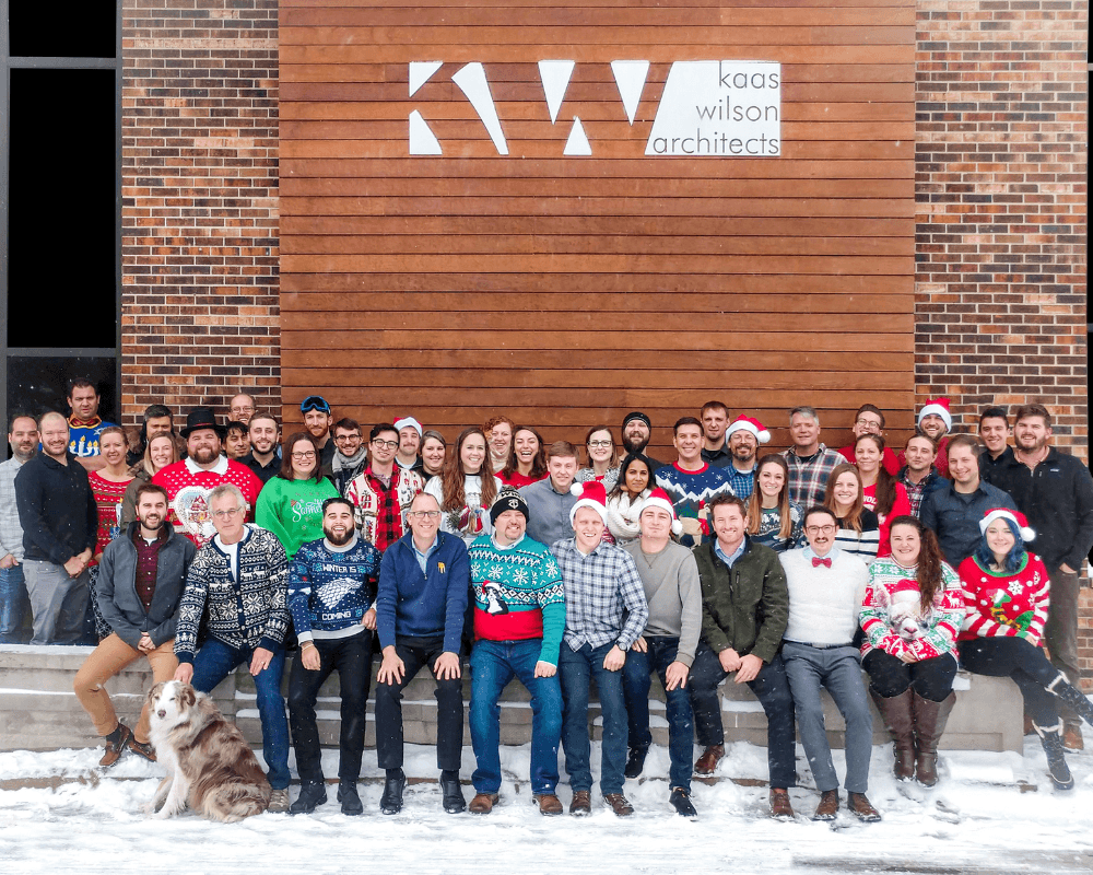 Large group photo of Kaas Wilson Architects team wearing holiday sweaters and winter attire, posed in front of their wood-paneled office entrance displaying the company logo. Team members are arranged in two rows on snowy ground, with a dog sitting at the far left. The brick building facade and company's geometric "KWA" logo are visible in the background.