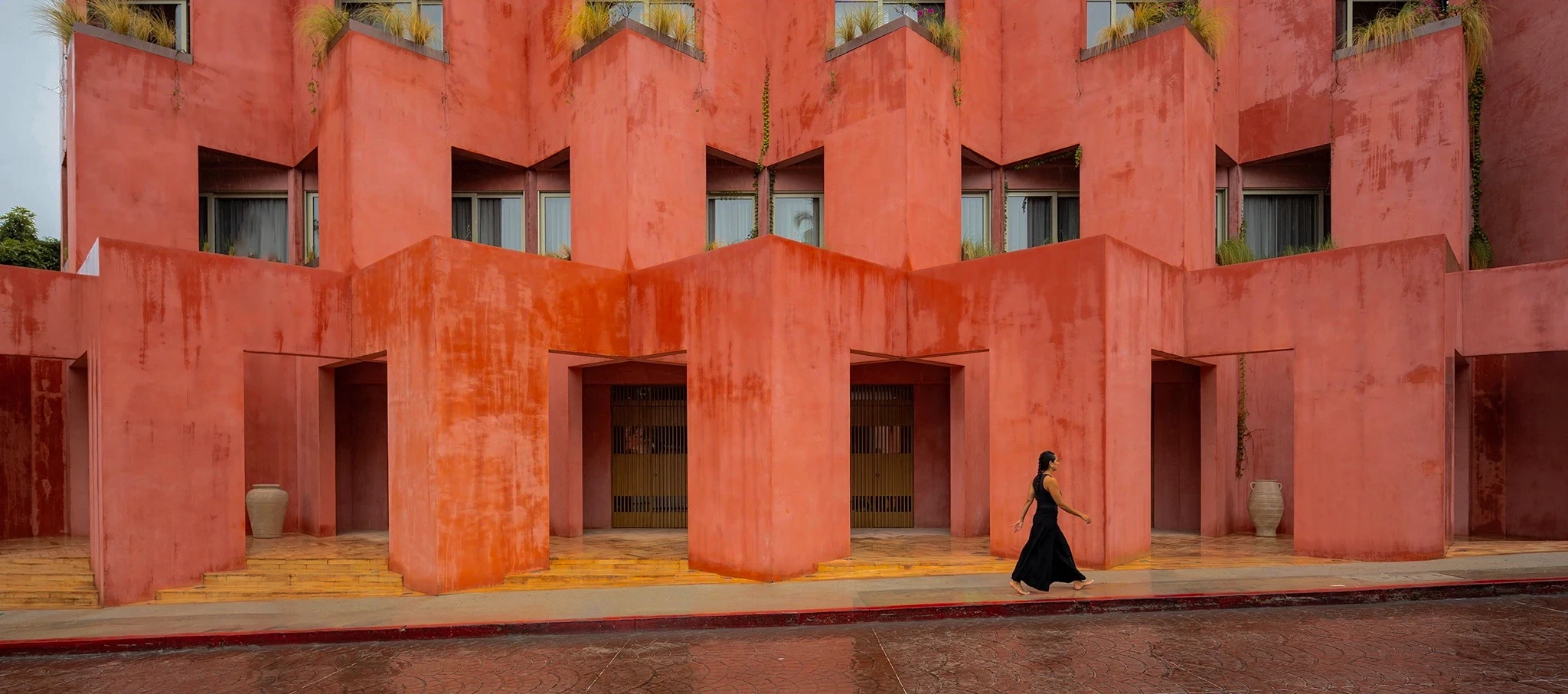 A woman in a black dress walking past a multi-storey terracotta red brutalist building with geometric protruding volumes