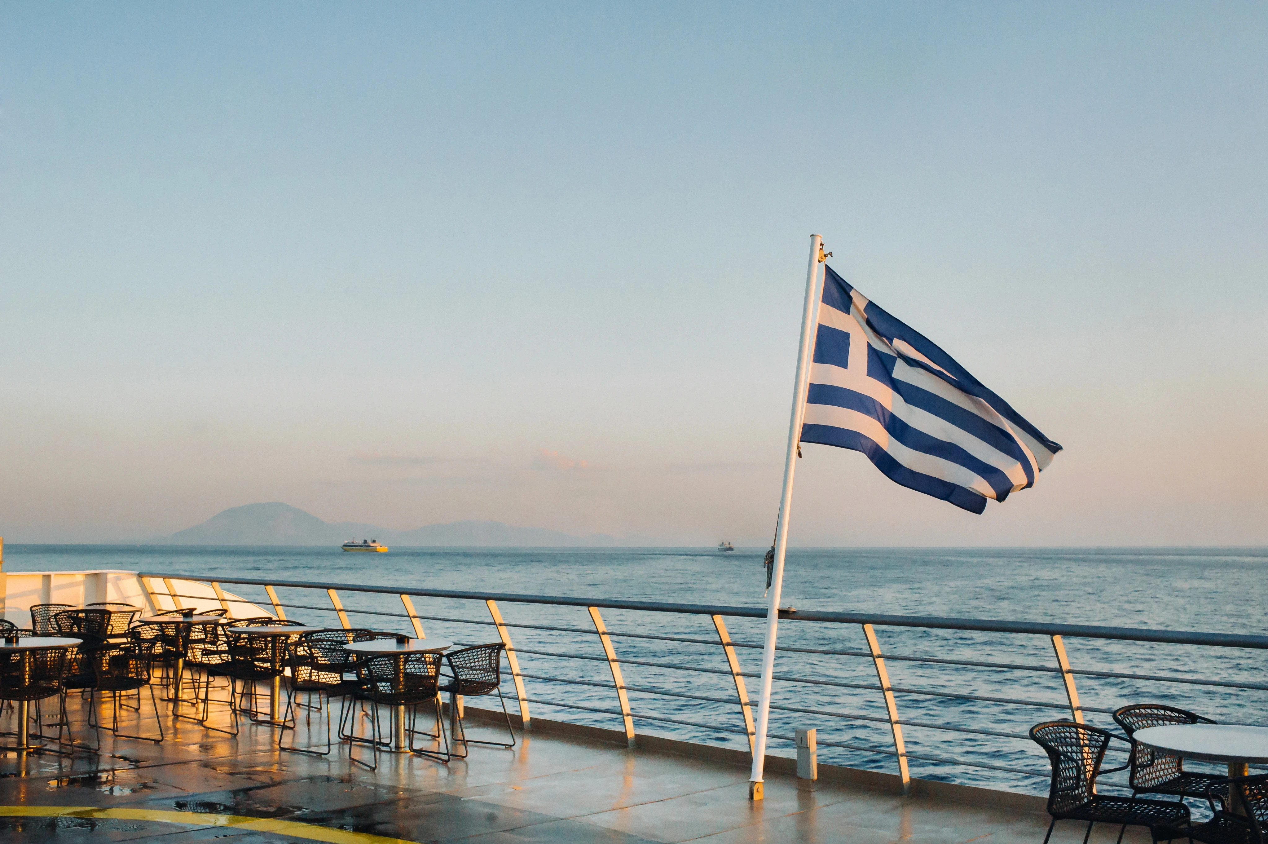A Greek flag waving on the deck of a ferry at sea during sunrise.