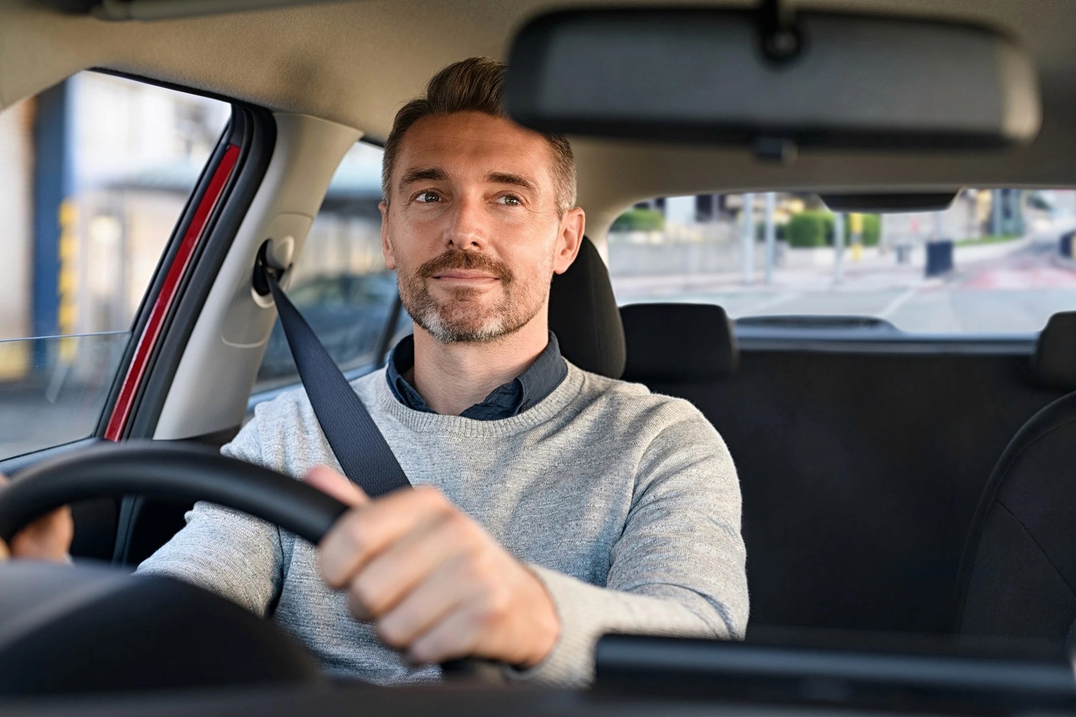A man driving a car. He is wearing a grey sweater and is looking at the camera with a slight smile.