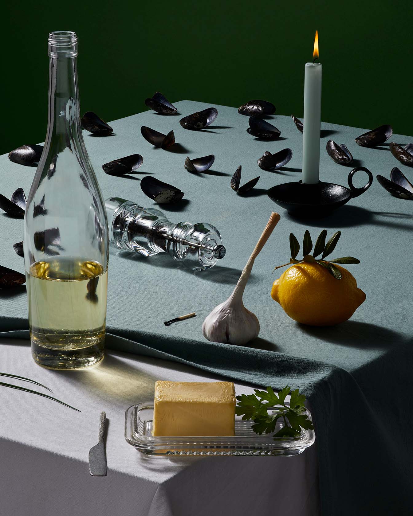 Ingredients including a bottle, a lit candle, garlic and a herb-topped dish, arranged on a printed cloth (dish unclear, please confirm with Michiel).
