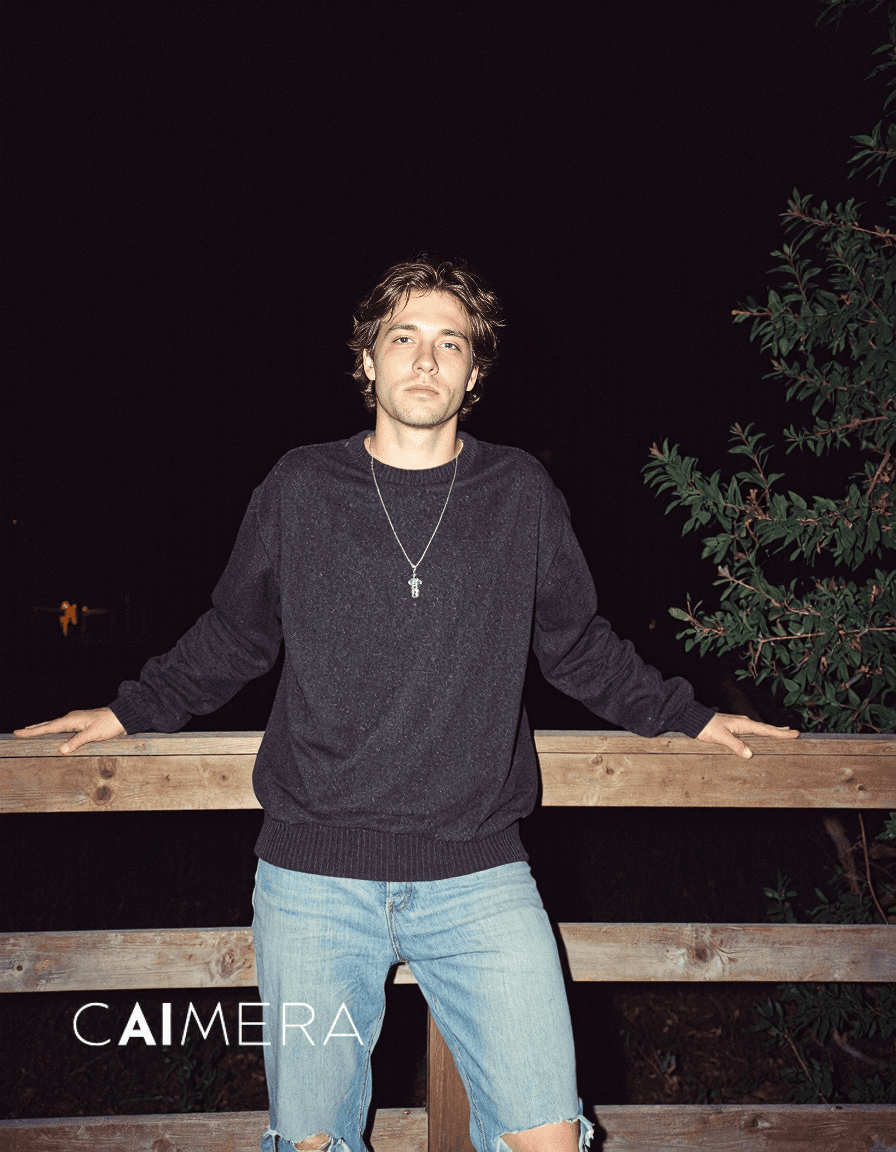 Moody nighttime portrait featuring person in dark sweater and light jeans leaning against wooden railing with foliage visible against dark sky