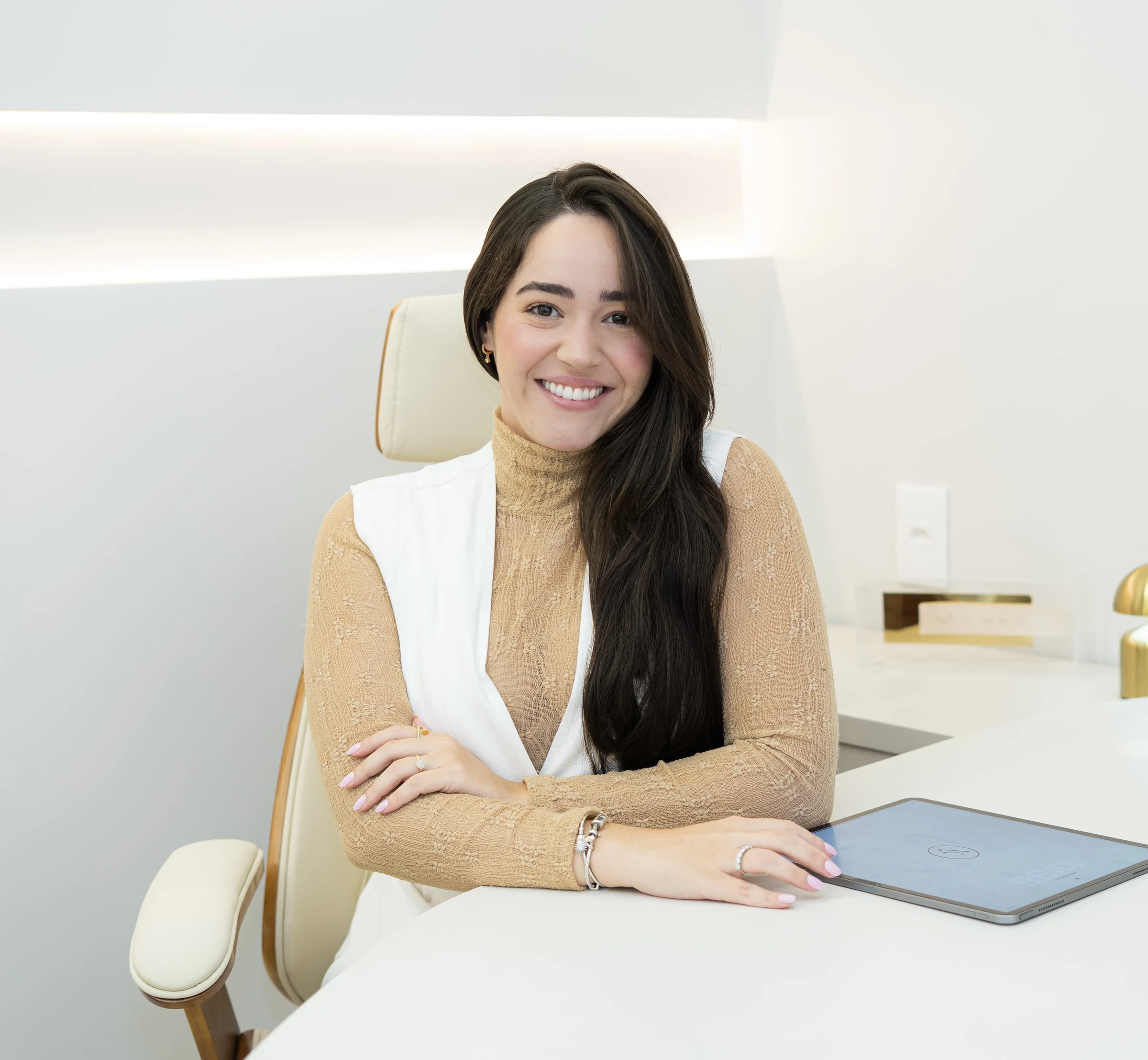 Doutora sorridente sentada à mesa branca, braços cruzados, com tablet ao lado