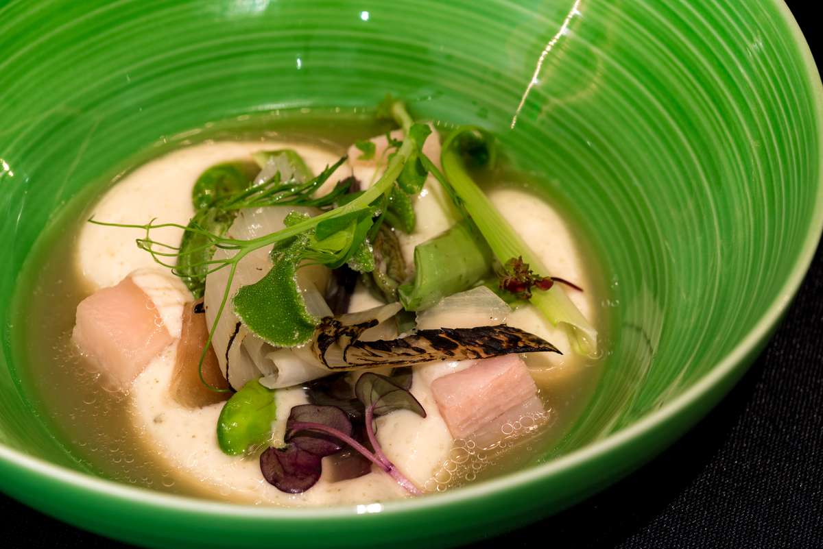 A green ceramic bowl contains a creamy soup with chunks of fish, green beans, herbs, and a grilled whole fish.