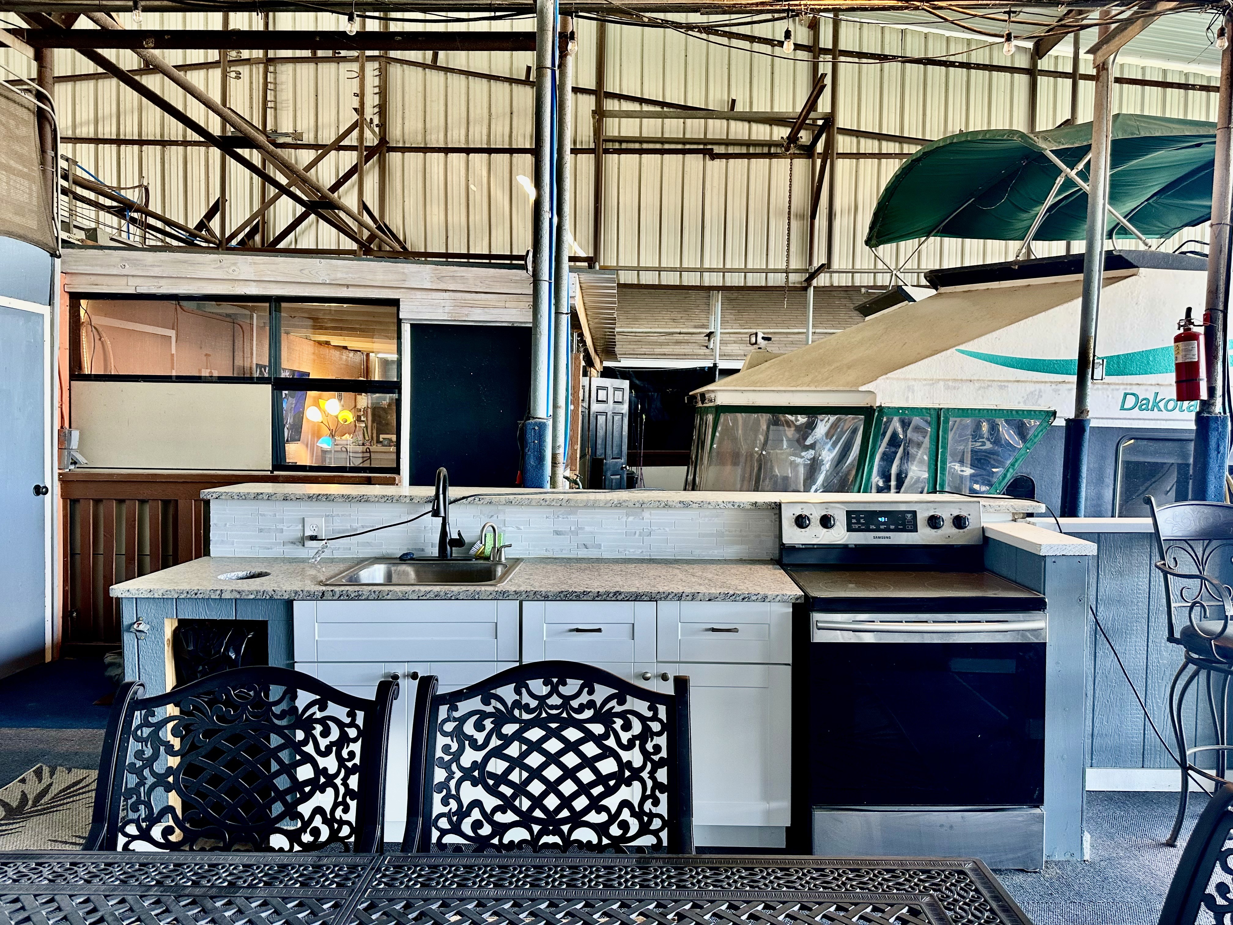 A spacious indoor boat storage facility features a modern outdoor kitchen setup with a sleek stovetop, sink, counter, and a patterned metal dining table, set against the backdrop of stored boats and an industrial-style ceiling.