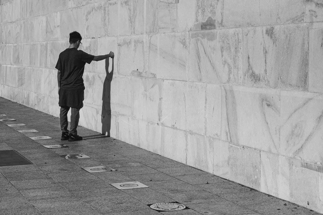 A man standing with one arm outstretched against a large marble wall, his shadow cast beside him on the pavement, black and white