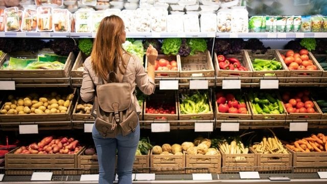 Mulher observando seção de hortifruti organizada, com legumes e verduras separados por categoria, em um mercadinho.
