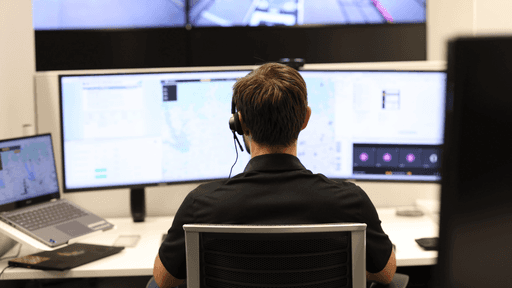 A person wearing headphones sits at a desk, analyzing data on multiple computer monitors displaying maps and diagrams in a modern control room environment.