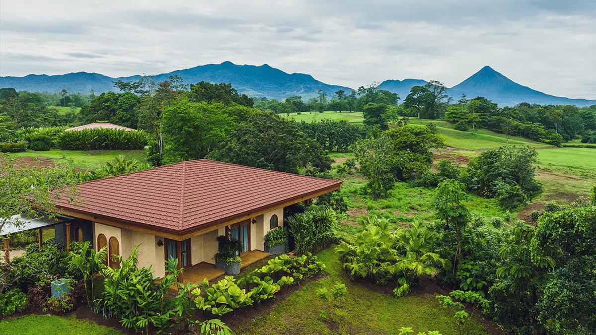 A single-story villa with a tile roof set in rainforest near La Fortuna, with Arenal Volcano and mountains behind under a cloudy sky.