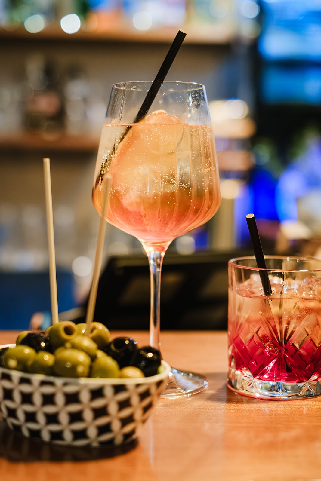 A chef wearing a striped apron, light-coloured shirt and cap speaks to diners while gesturing with his hands. He stands in a restaurant setting with glassware and a water carafe on the table in front of him, and shelves of ingredients and packaging visible in the background.