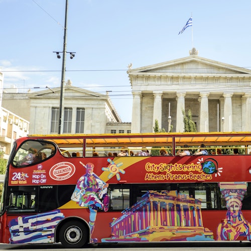 一辆色彩鲜艳的双层观光巴士，车身绘有雅典地标，从古典希腊建筑前驶过。