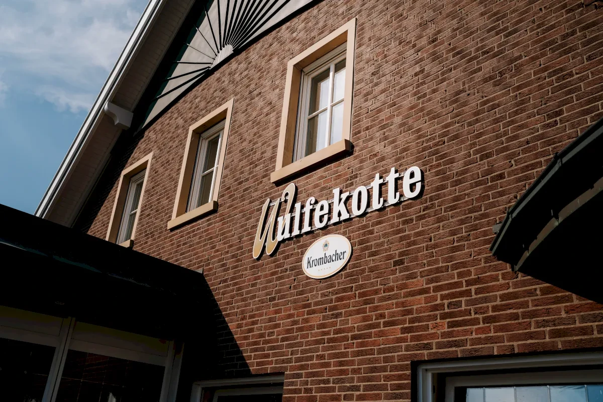 Das Schild "Hilfekotte" an der Backsteinfassade eines Gebäudes mit drei Fenstern, darunter ein Logo.