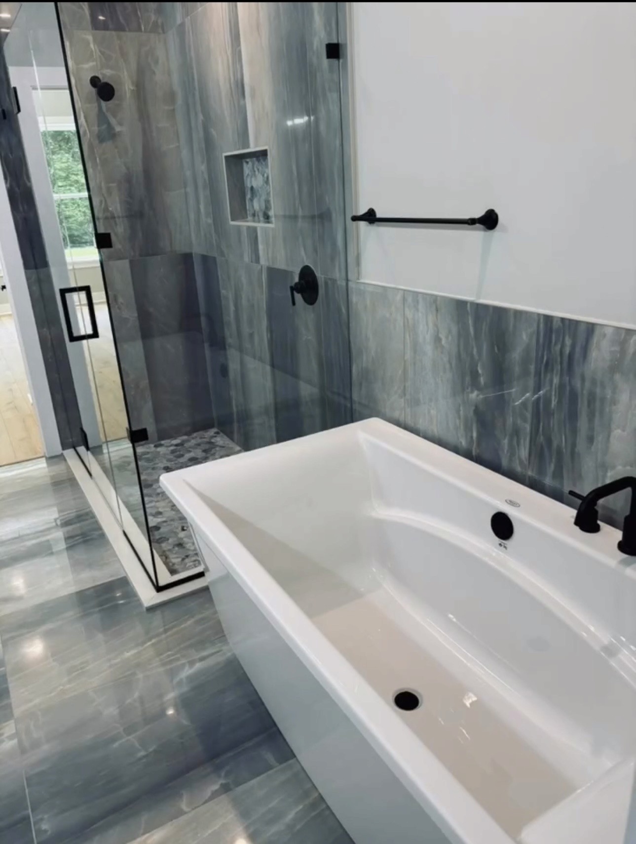 Master bathroom full view with double vanity, glass shower, soaking tub, polished gray marble floors, round black mirror, and natural light
