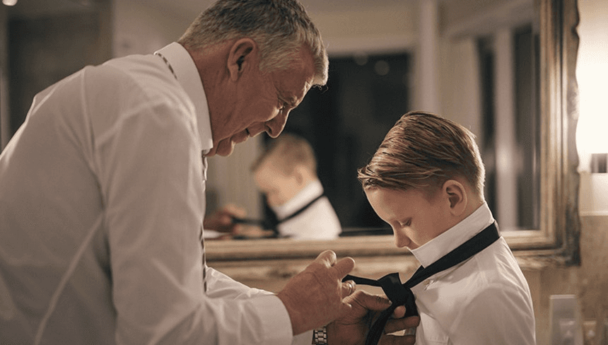 An older man helps a young boy adjust a bow tie in a warmly lit room, reflecting a moment of guidance and care.