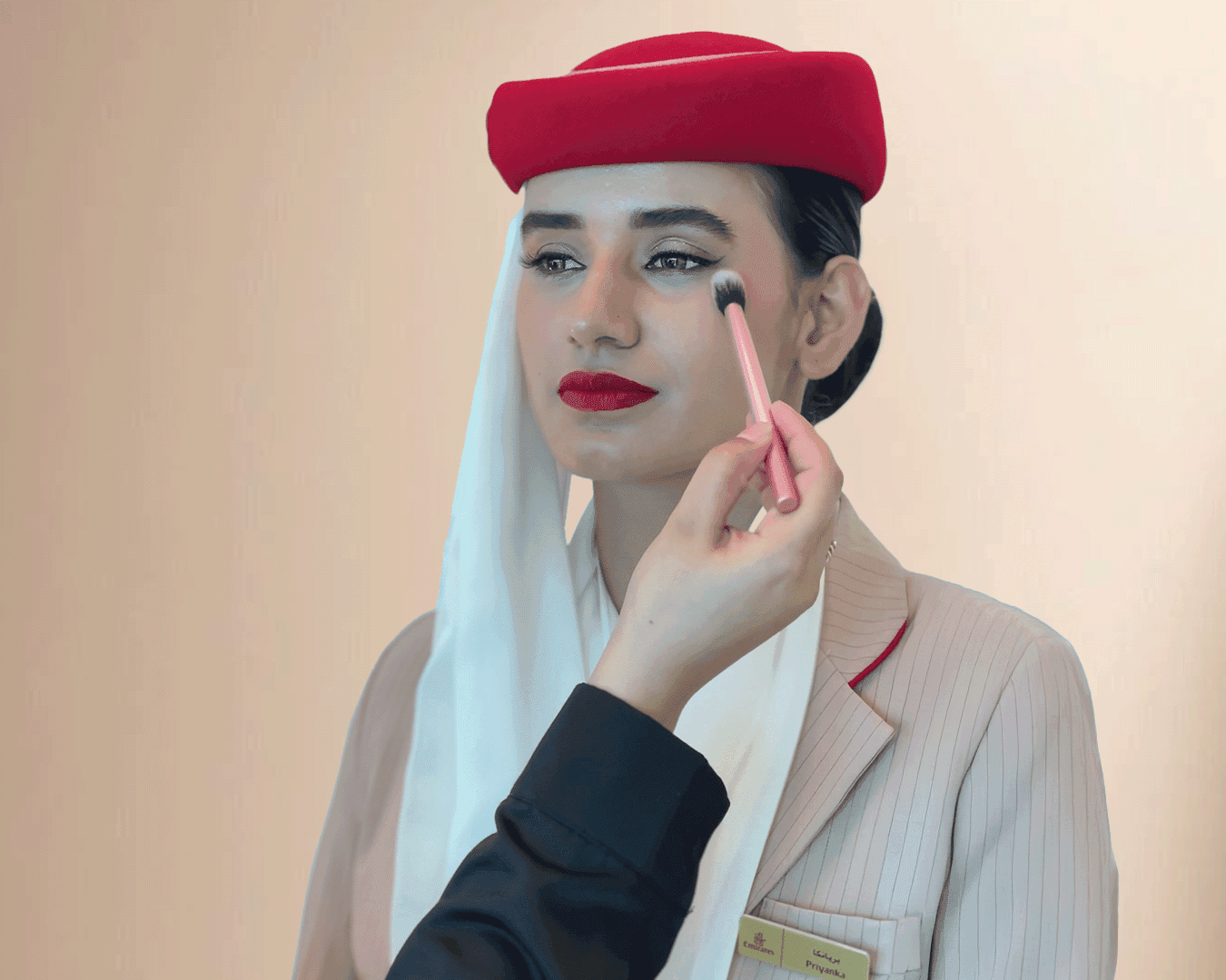 Woman in a graduation cap and gown receiving professional makeup, with a makeup artist applying blush to her cheek using a brush.