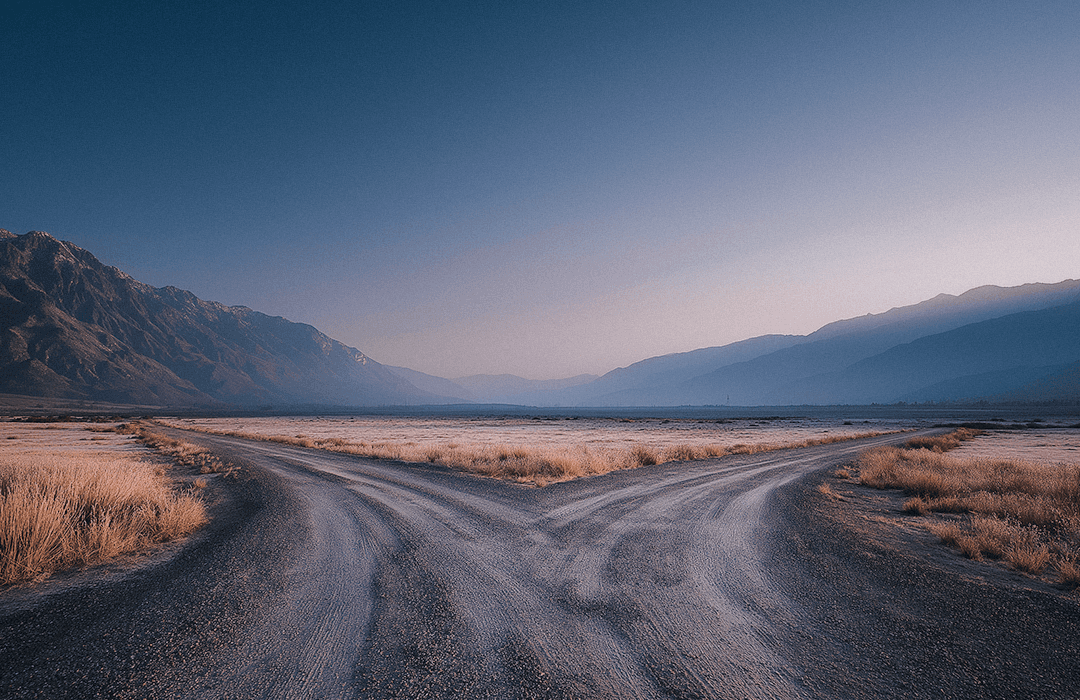 Uma estrada de terra que se divide em duas direções opostas em um cenário desértico, cercada por montanhas sob um céu claro ao amanhecer.