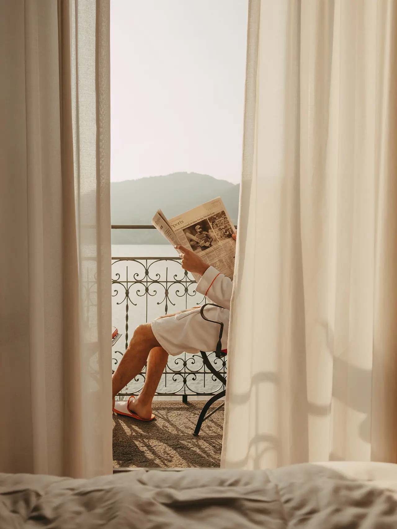 A person in a bathrobe sitting on a balcony reading a newspaper, with a scenic view of mountains in the background through sheer curtains.
