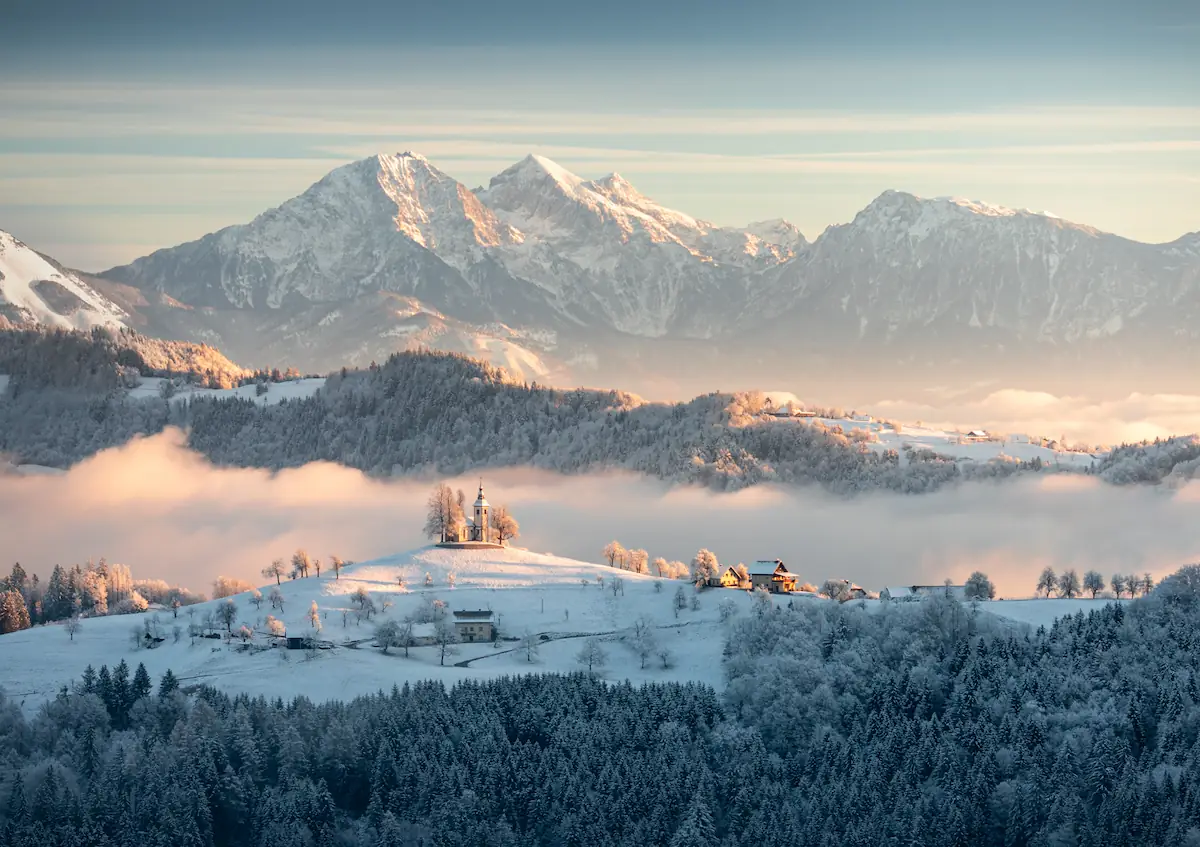 A wide winter landscape of Sveti Tomaž, Slovenia, featuring a hilltop church and scattered houses overlooking a valley of frozen trees under a soft dawn sky.