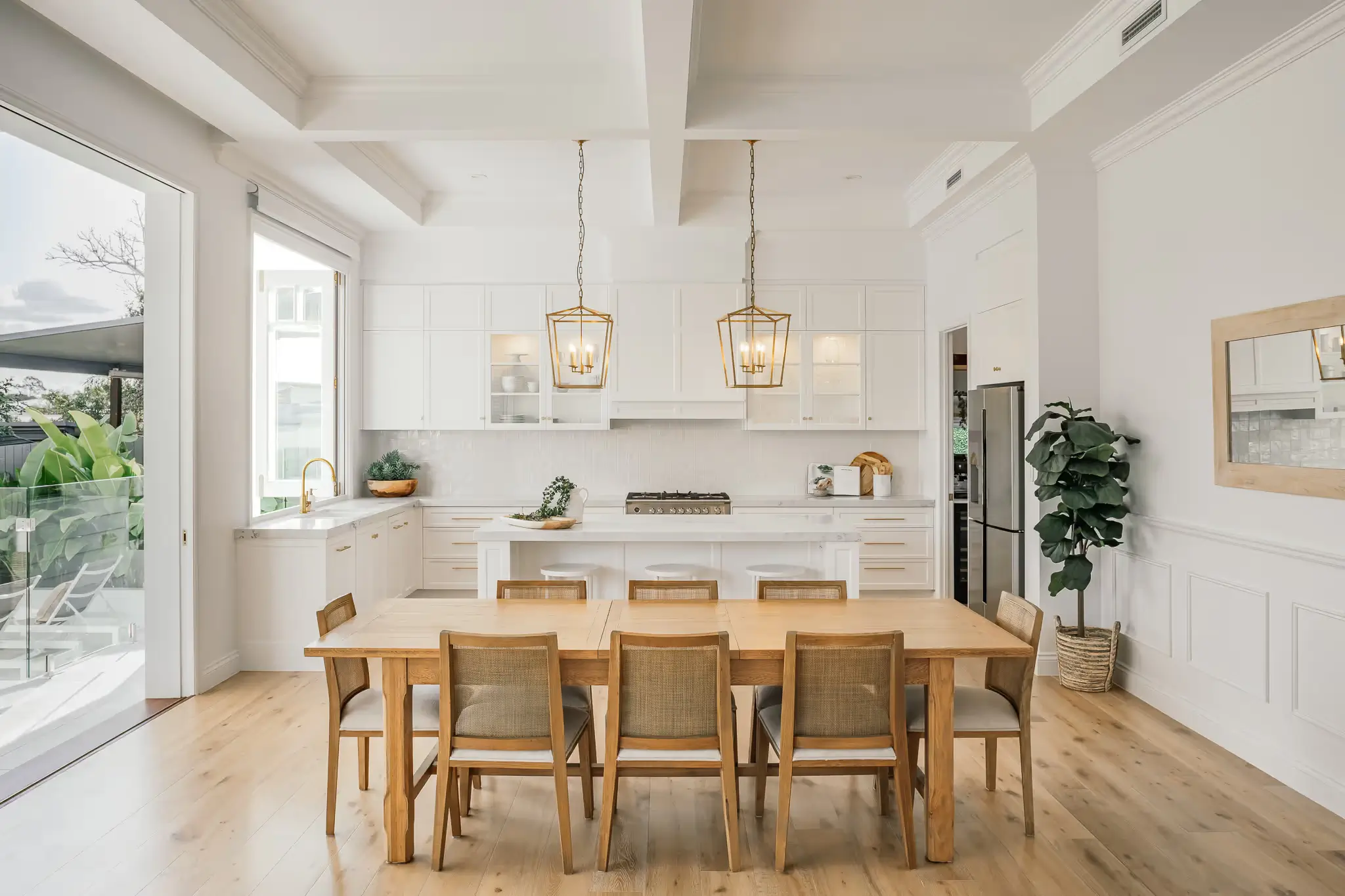 A dining table and chairs with kitchen in the backgriund with 2 pendants hanging from the coffered ceilings