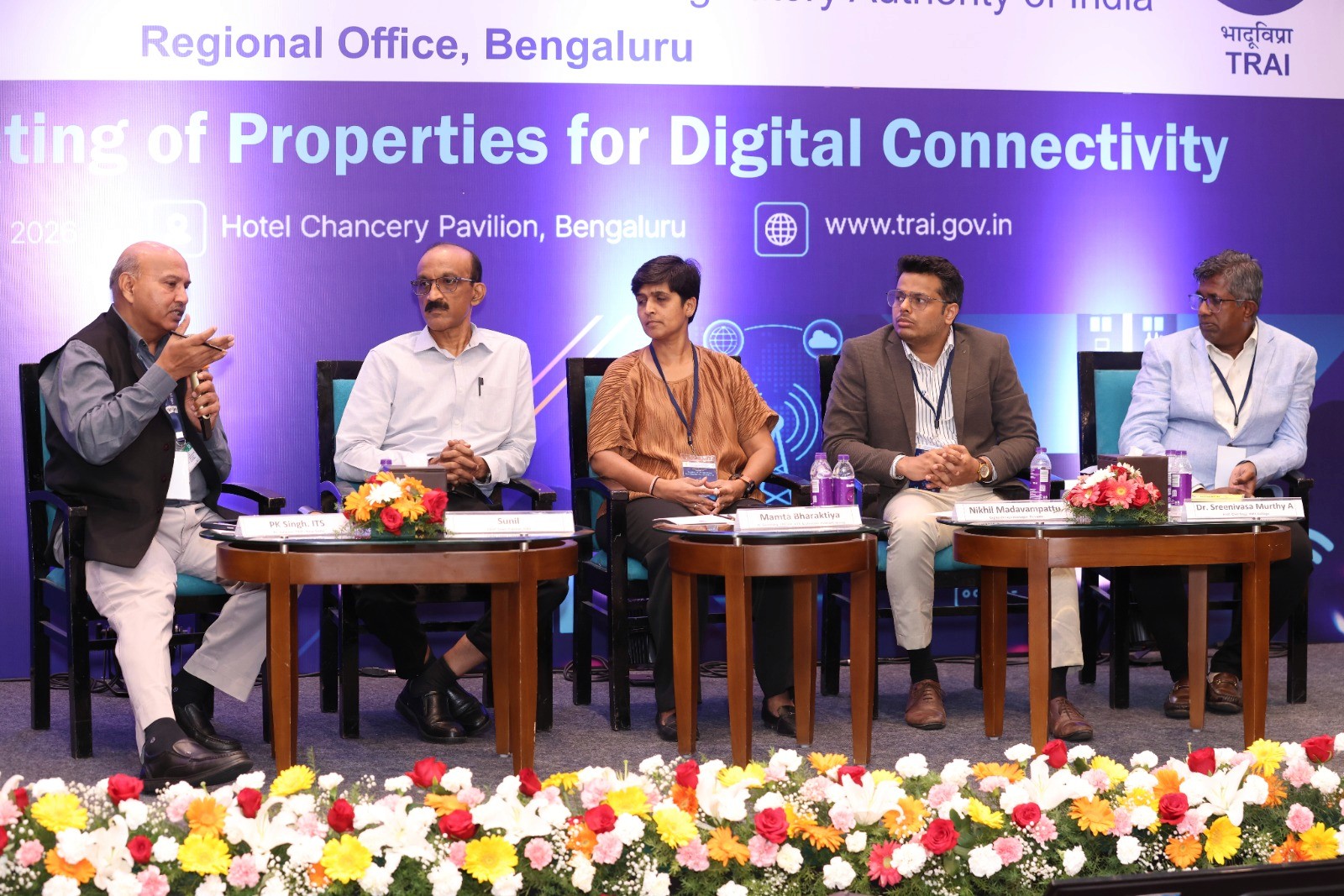A man stands while two people sit at a table in an office setting, smiling and engaged in conversation. The mood is friendly and collaborative.