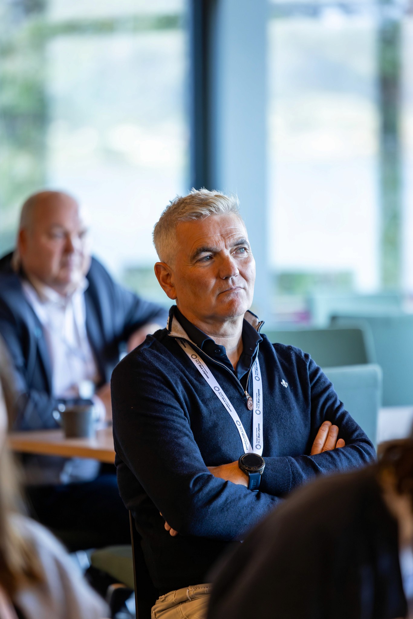 A man in a dark sweater sits in a conference setting, listening attentively with others in the background.
