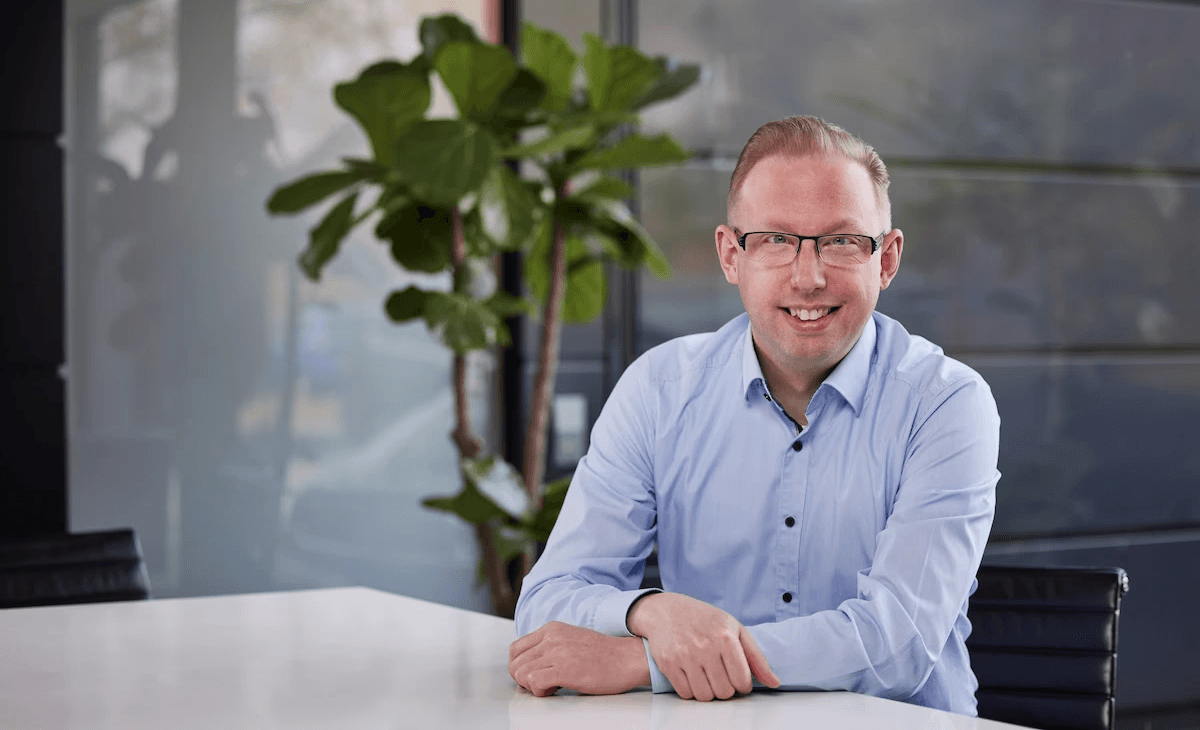 Person mit Brille und hellem Hemd sitzt an einem Tisch im Büro und lächelt, im Hintergrund eine Pflanze