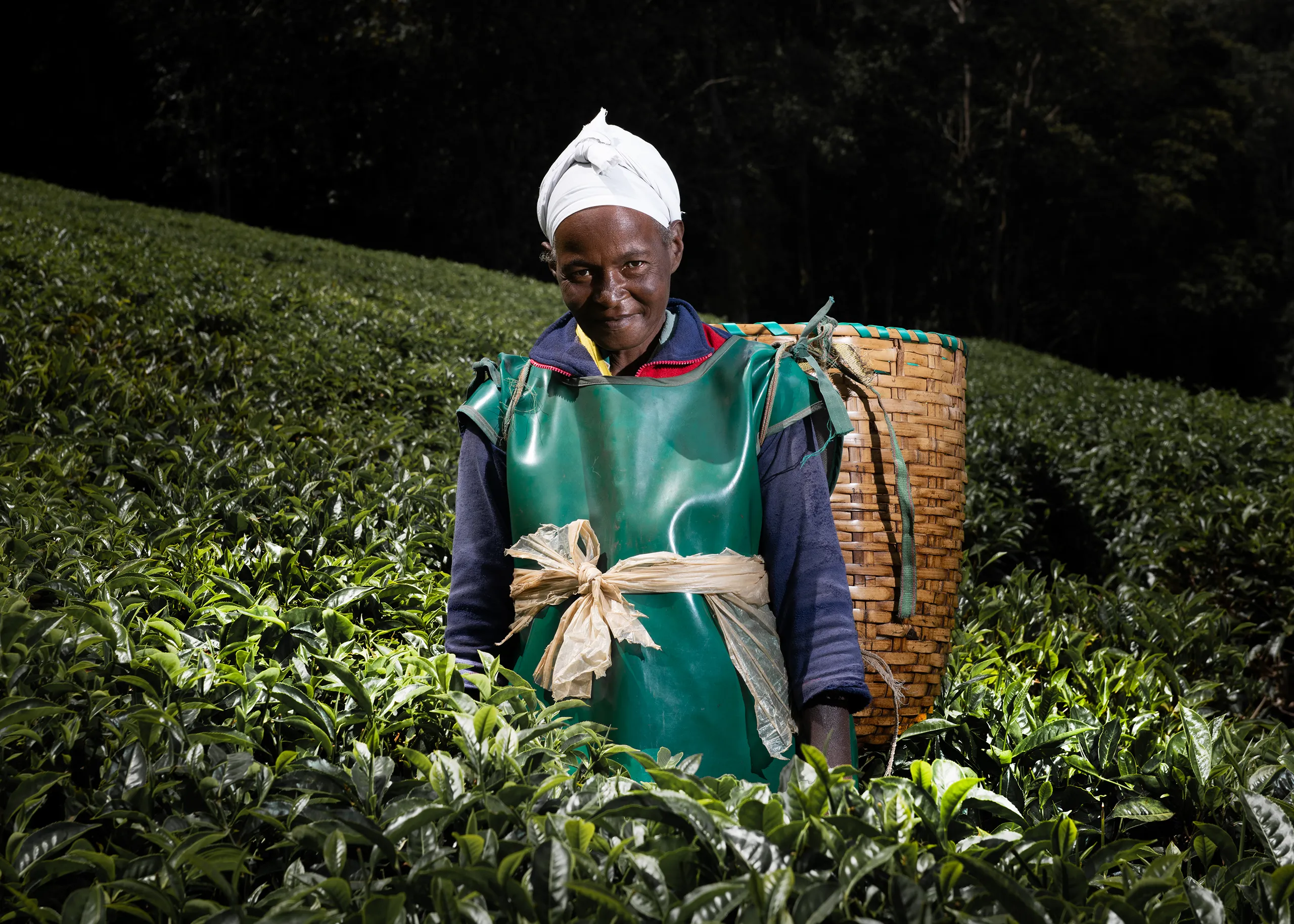 Editorial portrait of a female Kenyan coffee farmer with a white head wrap wearing a traditional collection basket.