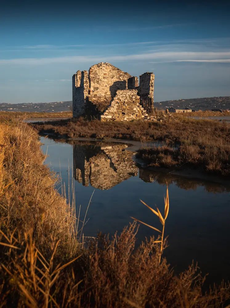 Historic stone ruins at the Sečovlje salt pans in Slovenia, mirrored in calm water with a coastal landscape in the distance.