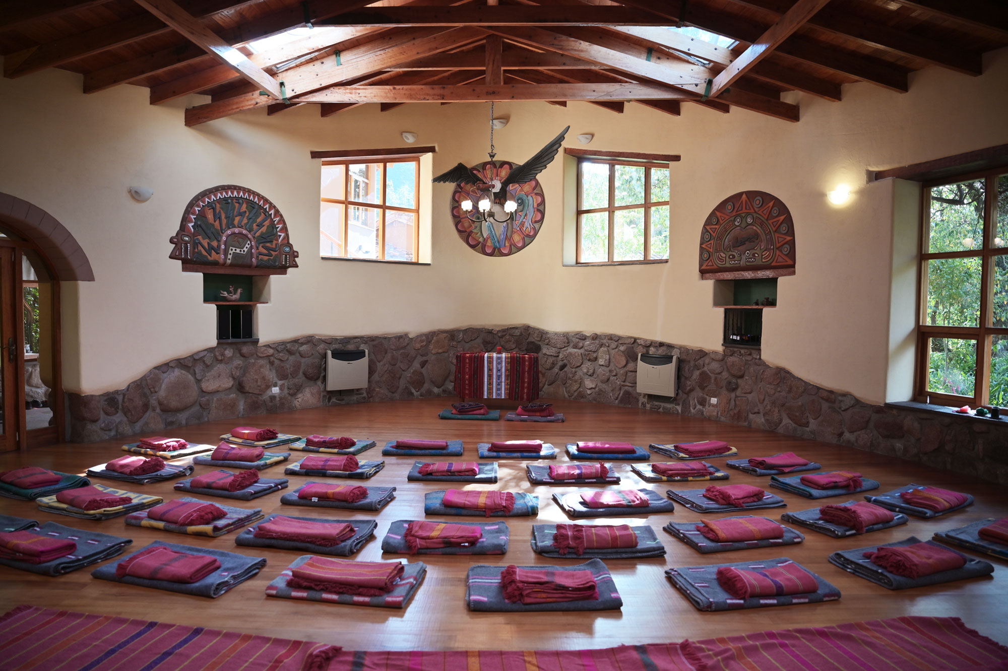 Spacious yoga room with wooden beams, decorative wall art, and large windows. Numerous mats and blankets are arranged on a wooden floor, creating a calm and inviting atmosphere.