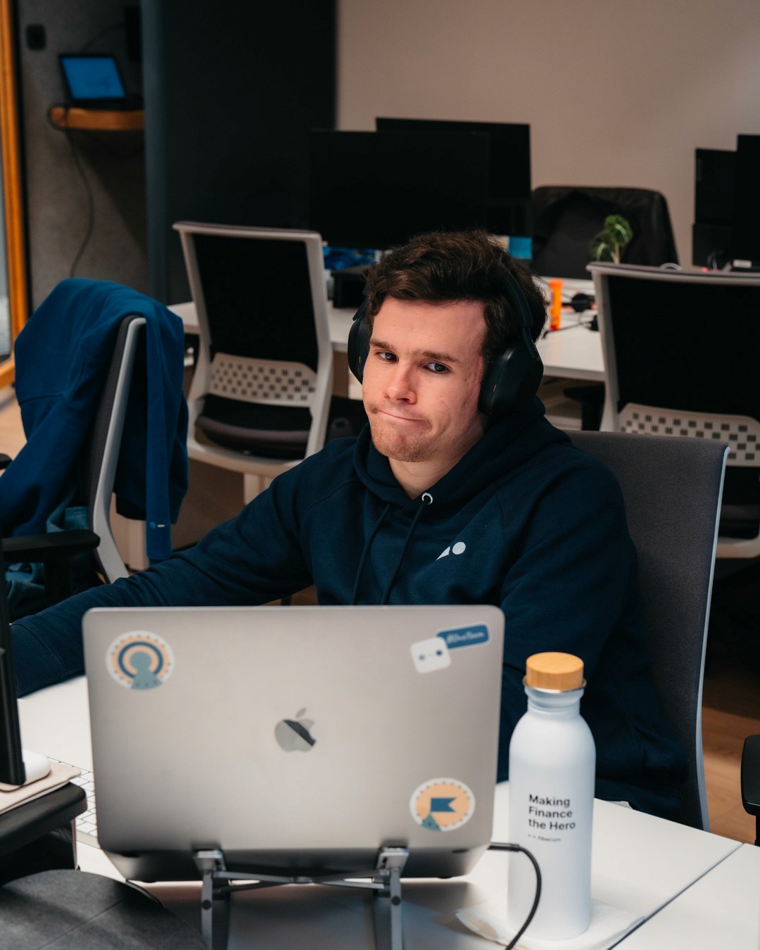 A young man wearing headphones sits at a desk with a laptop, focused on his work in a well-lit office space.