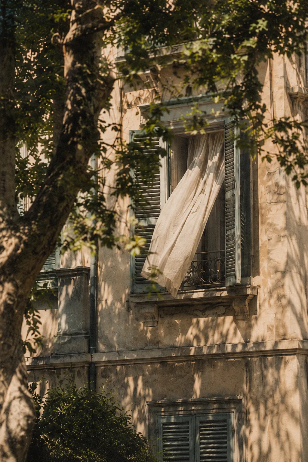 A white curtain blowing out of an open window with green shutters, on a pale stone building seen through tree branches.