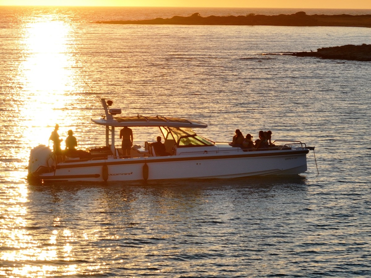 White Axopar 37 motor yacht anchored in calm waters at sunset near rocky Cycladic coastline with distant hills.