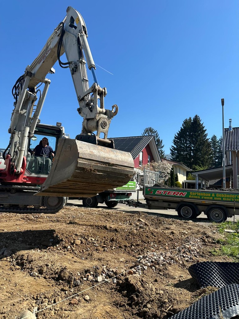 Bagger bei Erdarbeiten auf einer Baustelle vor einem Wohnhaus, daneben Anhänger mit der Aufschrift Gartenbau & Gebäudeservice.