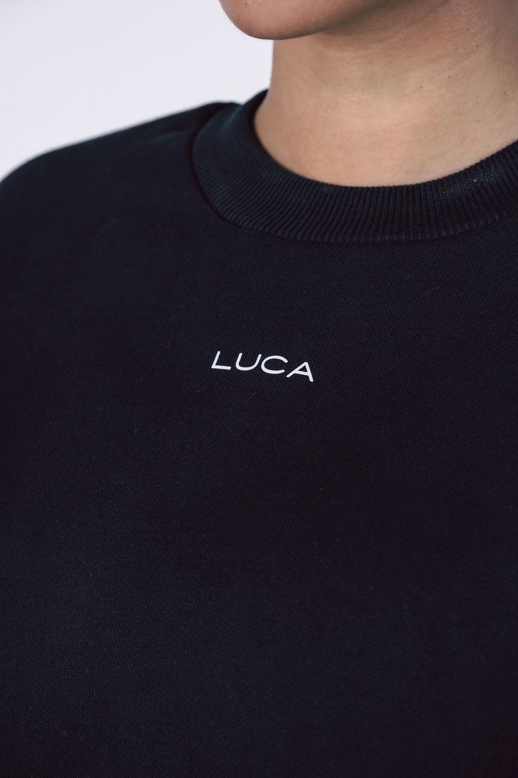 A close-up-detail-shot-of-a-black- crewneck-sweatshirt-showing-the- white-embroidered-LUCA-brand-logo- centered-below-the-neckline-Focus-on-black-crewneck-sweater- embroidered-logo-apparel-unisex- casual-wear-and-simple-modern- sweatshirt