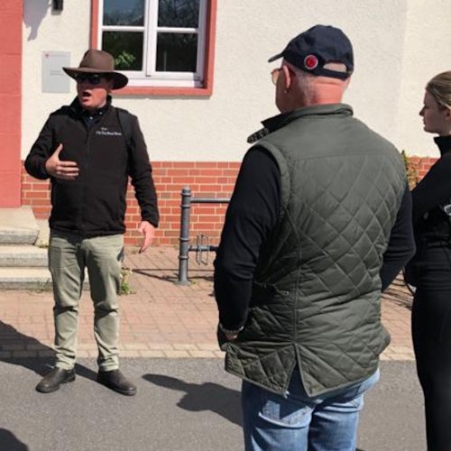 모자를 쓴 한 사람이 벽돌과 스터코 건물 근처 야외에 서 있는 두 명에게 이야기하며 손짓을 합니다.