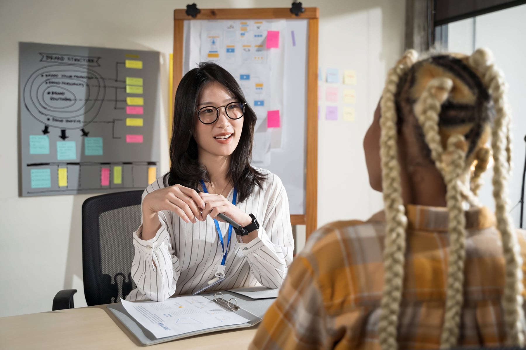 A woman wearing glasses sits at a desk during a meeting or interview, smiling and speaking with another person. Behind her are whiteboards covered in diagrams and colourful sticky notes.