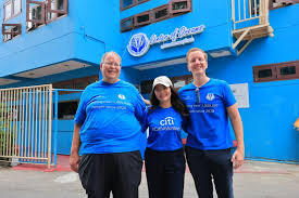 man in blue t-shirt and white floral skirt holding black smartphone while sitting on sidewalk
