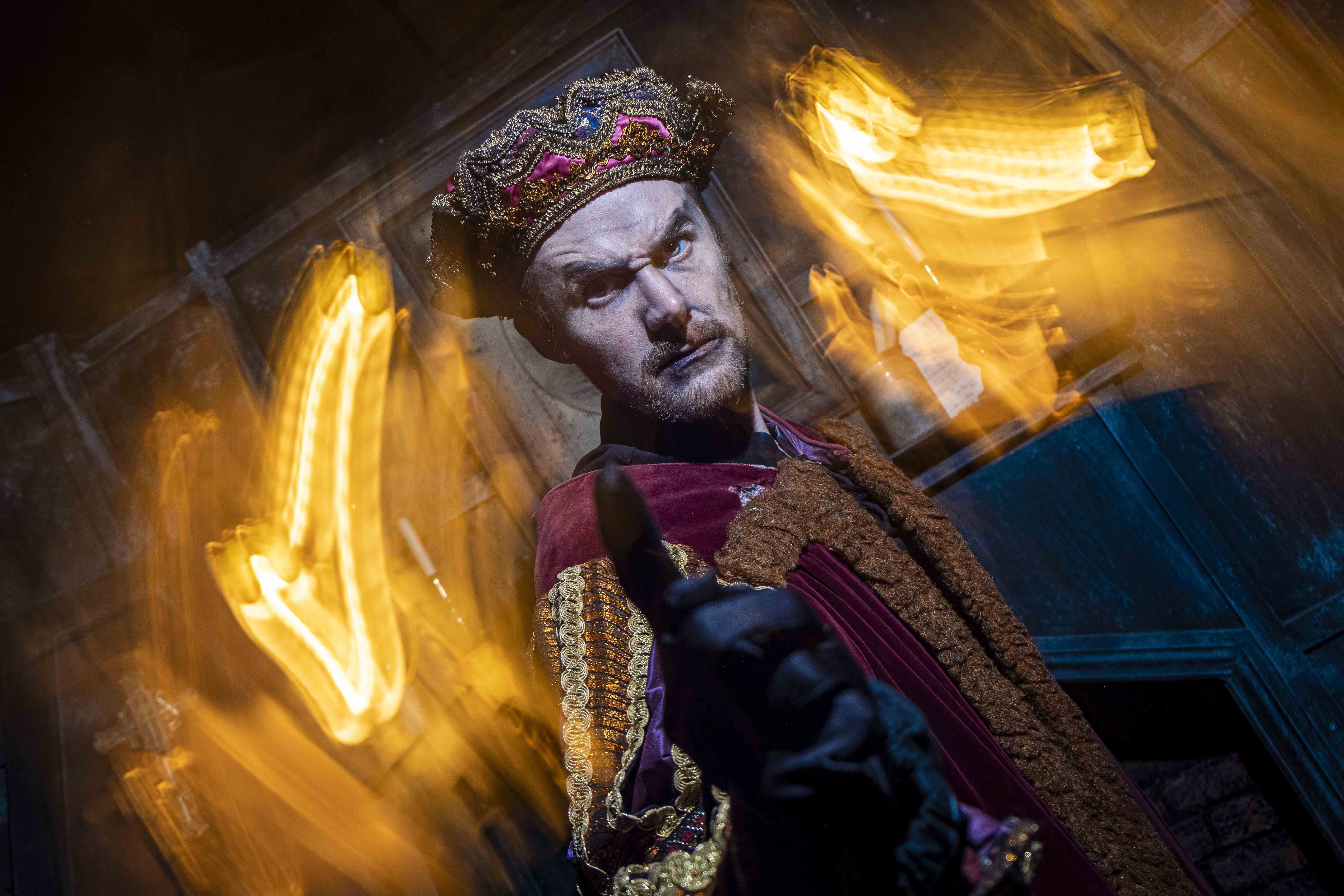 A person dressed in an ornate medieval costume with a richly decorated crown and a regal expression stands in a dimly lit room with blurred, glowing lights creating an intense atmosphere.