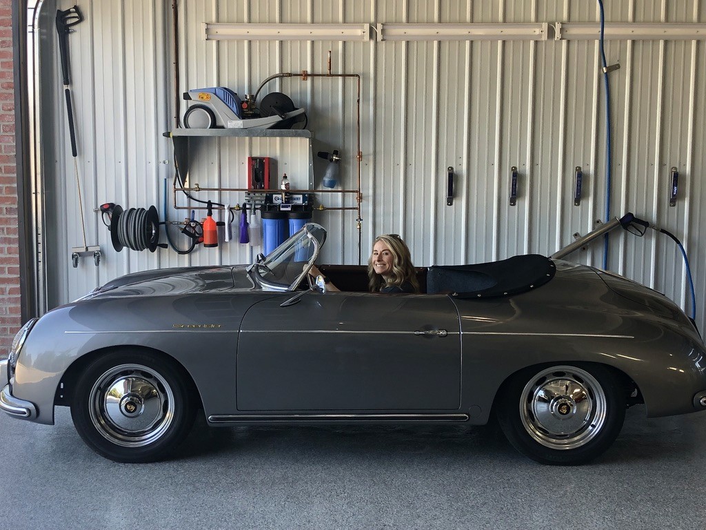 A vintage gray convertible car is parked inside a garage with a white corrugated metal wall, featuring various tools and a hose neatly organized on the wall.