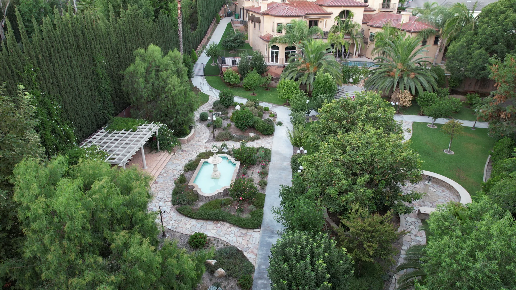 Garden paths surround a fountain, with planted beds and a home beyond.