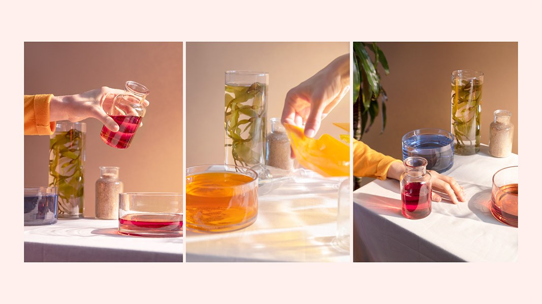 A triptych shows three sequential images of a person’s hands interacting with glassware containing colored liquids. The left image shows a hand holding a glass with red liquid and another with green liquid. The center image shows a hand placing a yellow leaf-shaped object into a tall glass. The right image shows a hand reaching toward a blue glass on a white tablecloth. Additional glassware and containers are visible on the table.