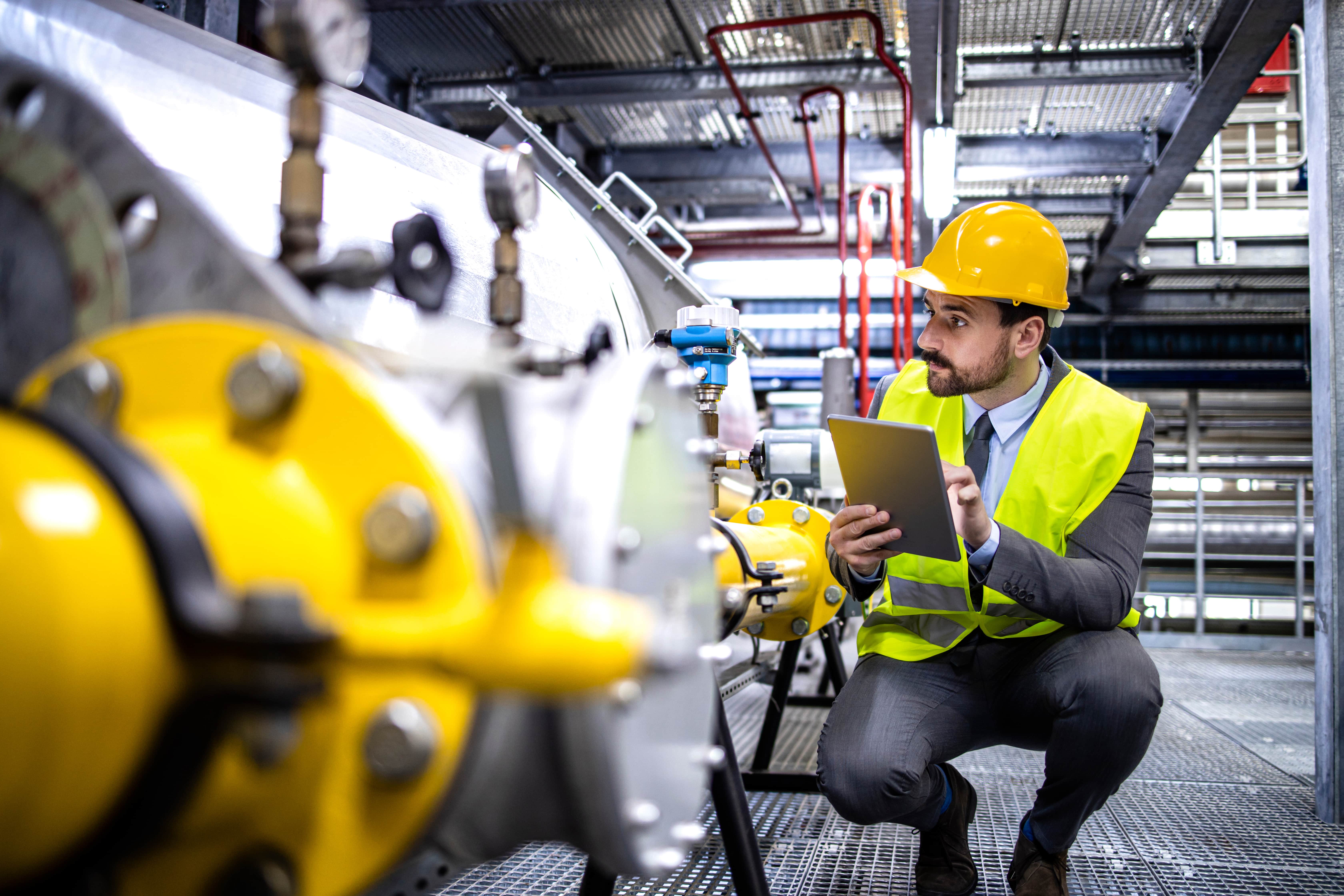 A male engineer wearing a hard hat and safety vest, inspecting industrial equipment with a tablet.