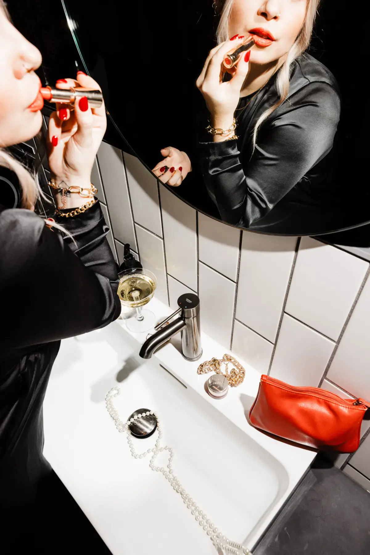 Woman applying red lipstick in a tiled bathroom mirror with jewellery on the sink.