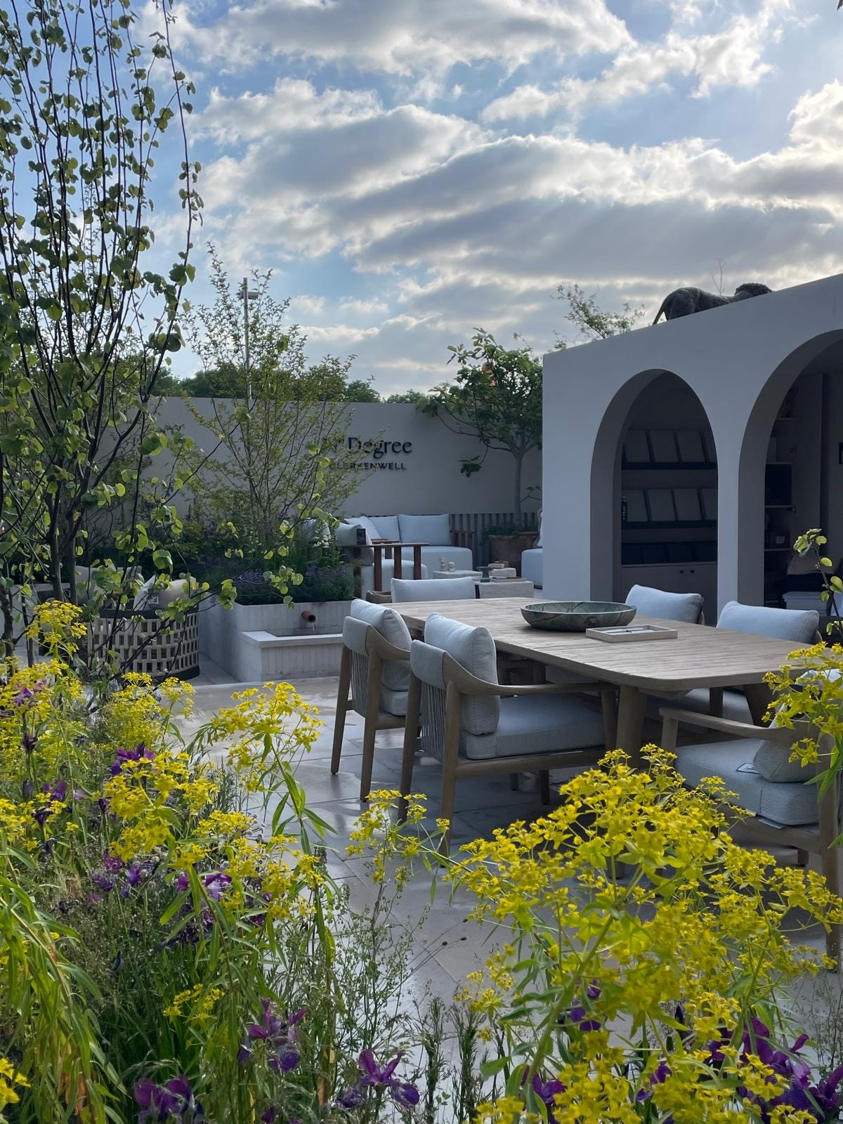A scenic view of a garden with flowers, a patio area, and clouds in the sky.