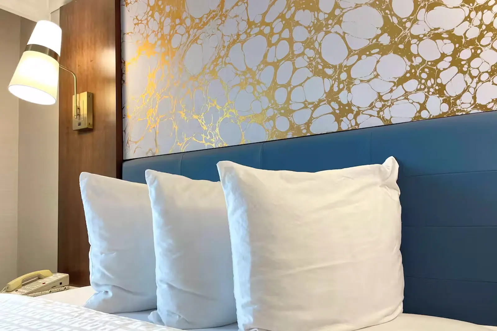 Three white pillows on a bed with a blue headboard and abstract gold wall art.
