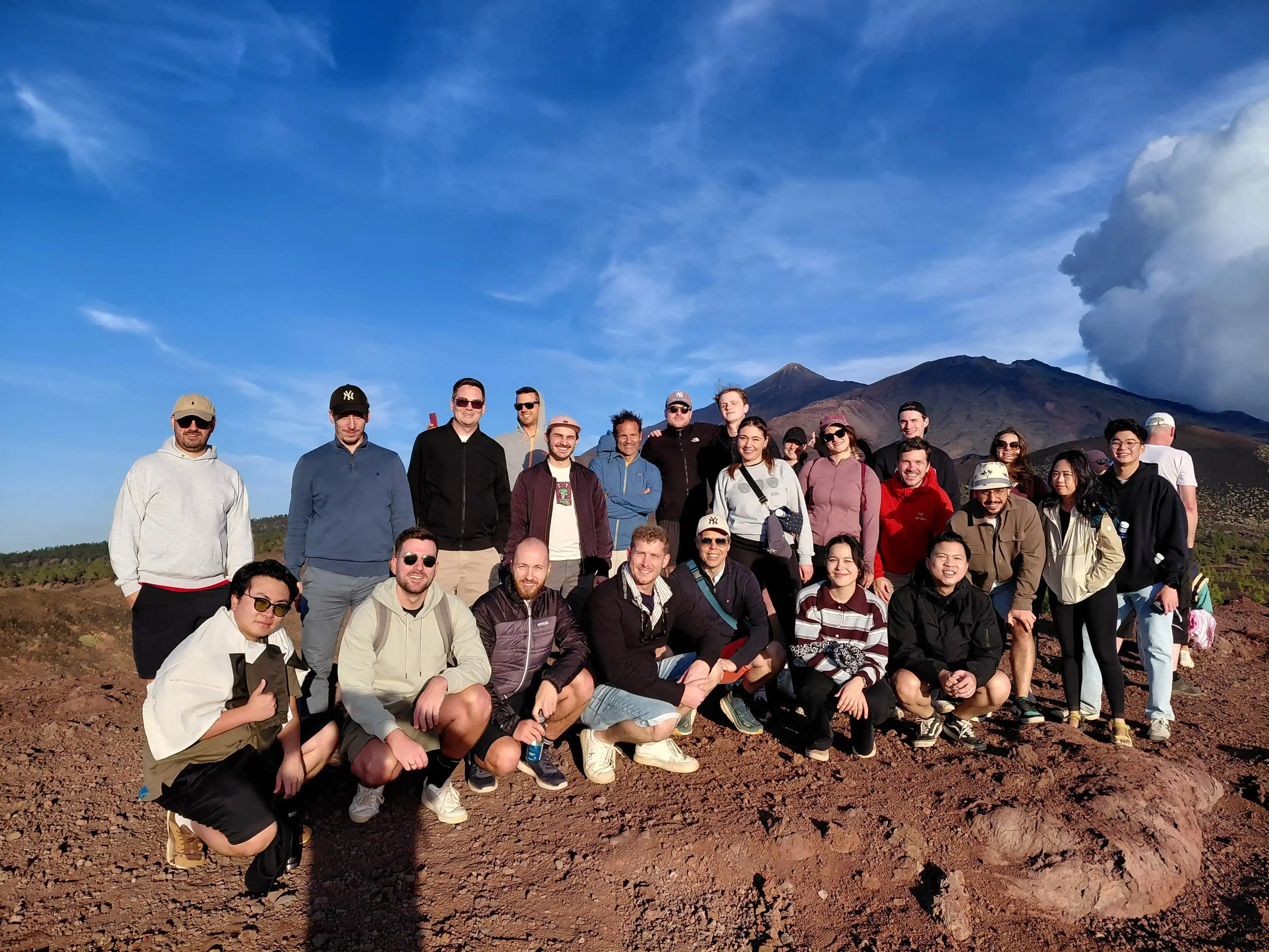 Large group photo outdoors on volcanic terrain with mountains and blue sky in the background