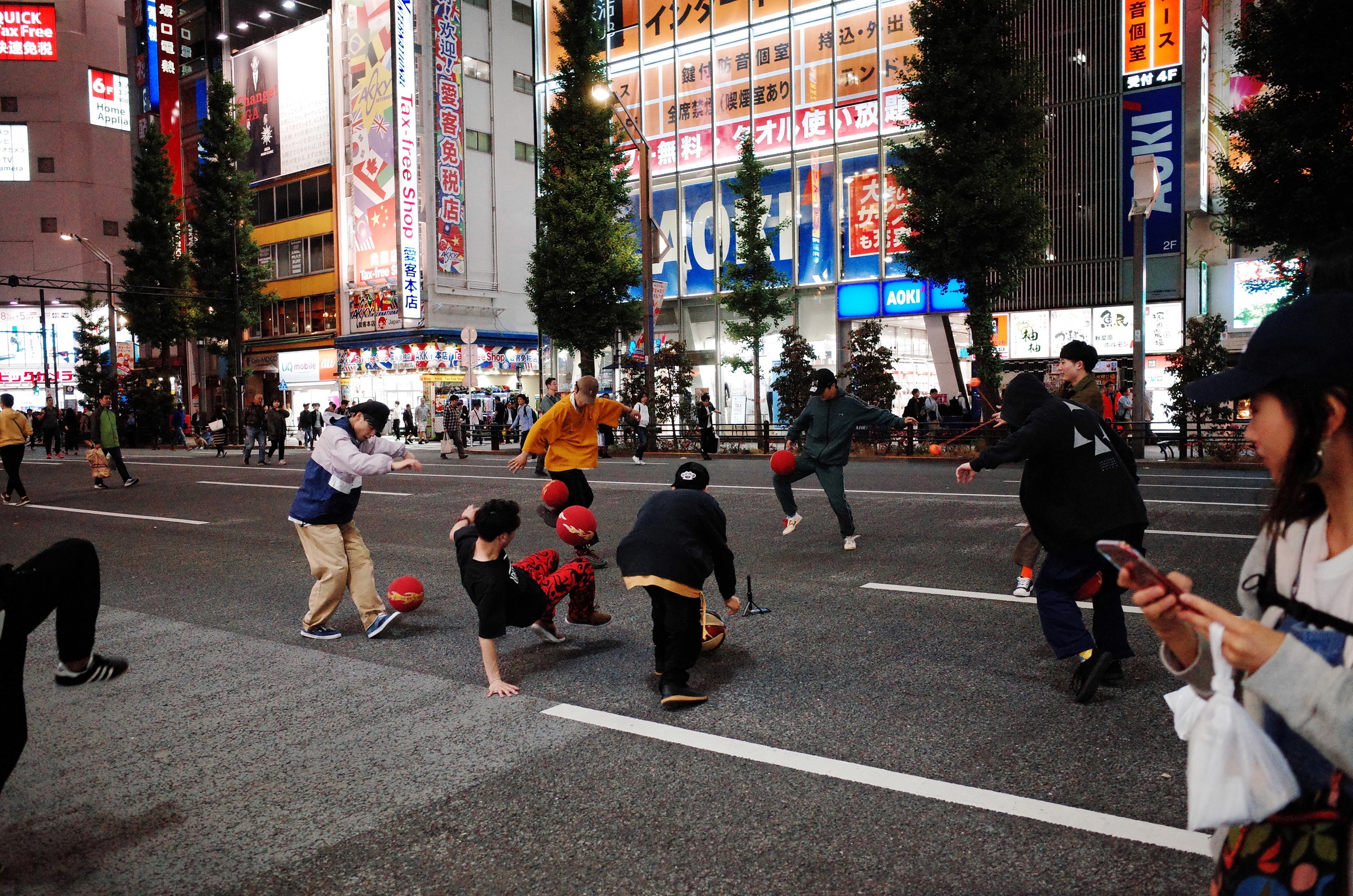 Photographie de rue prise la nuit en plein centre d'Akihabara montrant un groupe de jeunes en cercle qui jouent avec des balles et font du breakdance au milieu de la rue