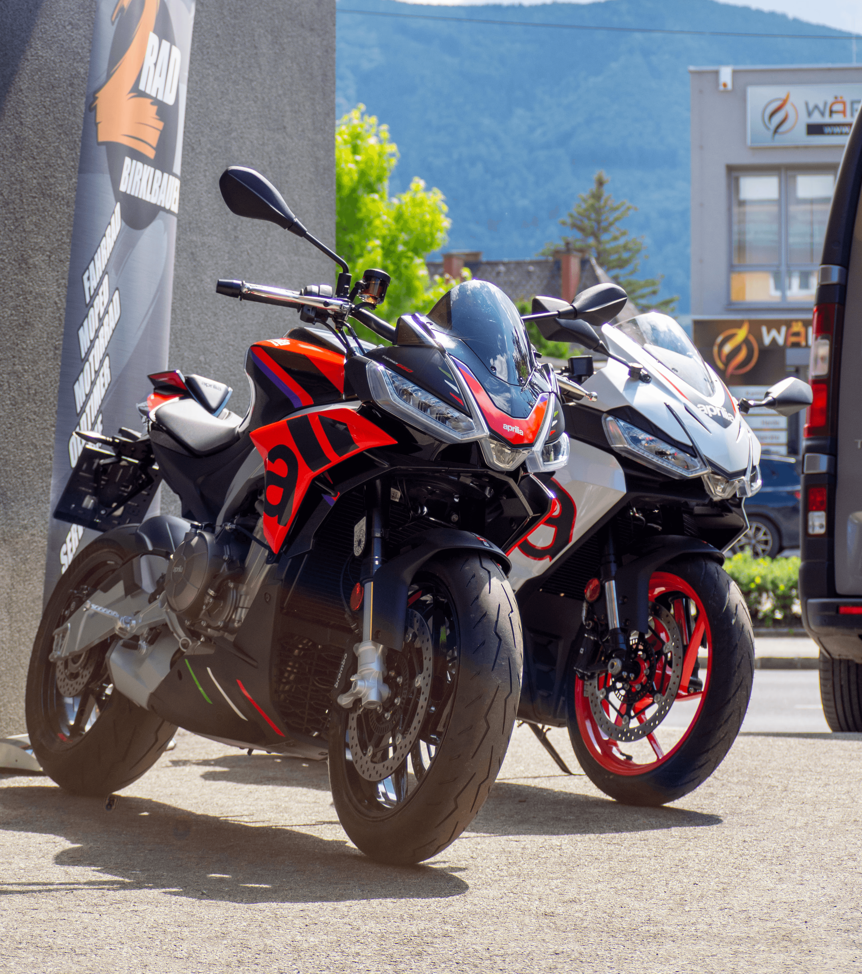 Two sporty motorcycles parked side by side, with a sunny backdrop and buildings in the distance.